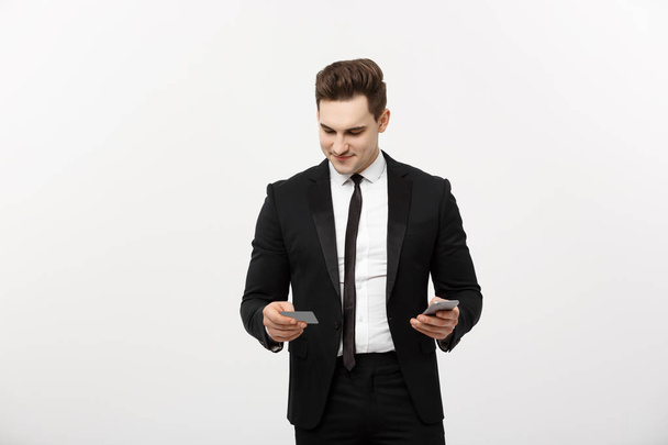 Портрет счастливого молодого бизнесмена в умном черном костюме, стоящего изолированно на фоне белой стены. Держать мобильный телефон и кредитную карту
. - Фото, изображение
