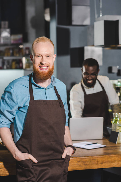smiling young male barista in apron with colleague in coffee shop  - Foto, imagen