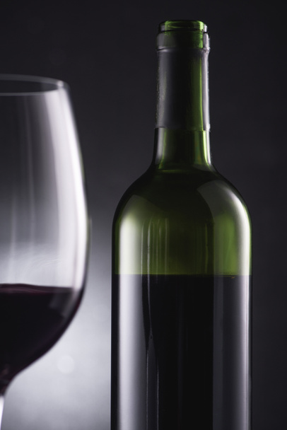 Detailní záběr rozmazané skla naplněné červeným vínem na popředí a víno láhev na černém pozadí - Fotografie, Obrázek