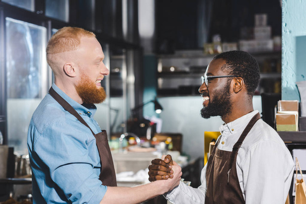 side view of two young multiethnic owners of coffee shop in aprons shaking hands in coffee shop  - Photo, Image