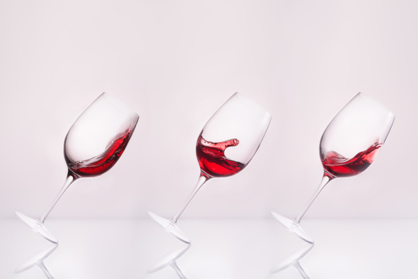 row of inclined wineglasses with splashing wine on reflective surface and on white - Foto, Imagem