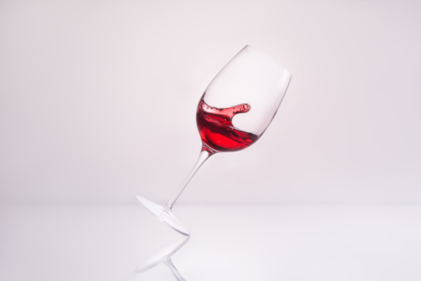 geneigtes Weinglas mit spritzendem köstlichen Rotwein auf reflektierender Oberfläche und auf weißem - Foto, Bild