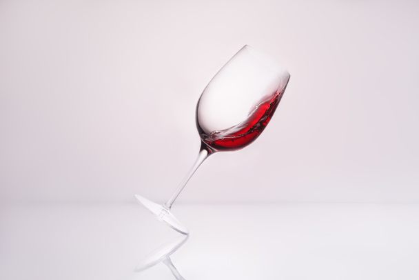 κεκλιμένη wineglass με κόκκινο κρασί στην αντανακλαστική επιφάνεια και σε λευκό - Φωτογραφία, εικόνα