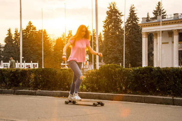 A young woman is riding a skateboard in a park in summer - Фото, изображение