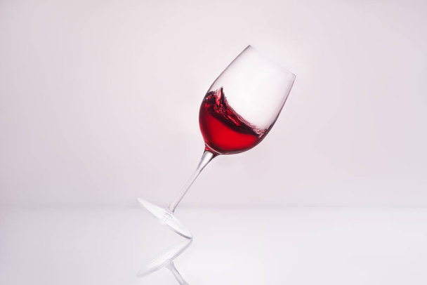 inclined wineglass with splashing red wine on reflective surface and on white - Foto, Imagem