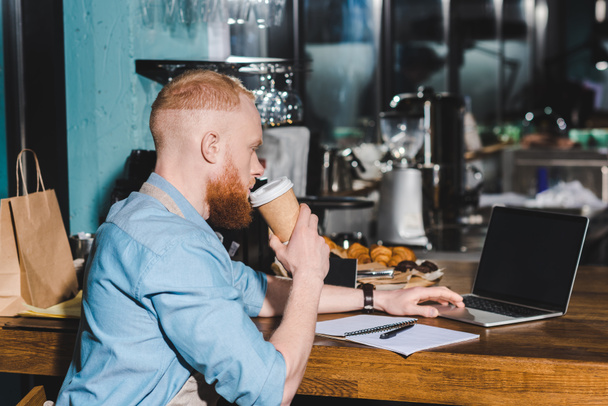 boční pohled mladých mužů barista pití kávy z papírového kelímku a pomocí přenosného počítače  - Fotografie, Obrázek