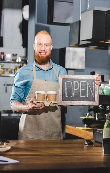 joven macho barista sosteniendo pizarra con letras abiertas y dos papeles tazas de café
  - Foto, Imagen