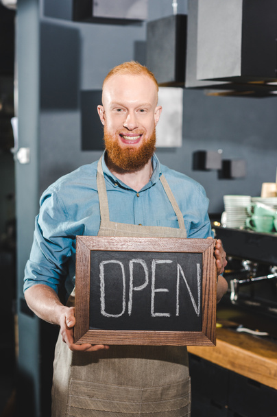 smiling young male barista holding chalkboard with lettering open in coffee shop - Photo, Image