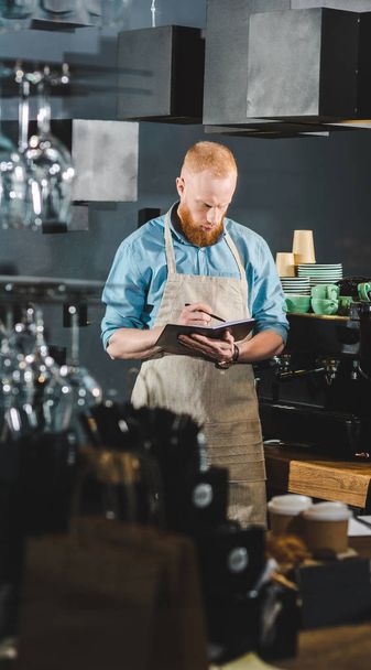 front view of young male barista in apron writing in textbook  - Foto, immagini
