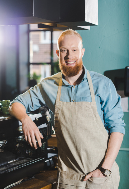 young bearded smiling male barista in apron standing near coffee machine  - Zdjęcie, obraz