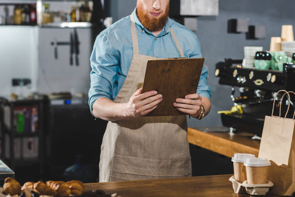 oříznuté shot z vážných mladý vousatý barista drží schránky v kavárně - Fotografie, Obrázek