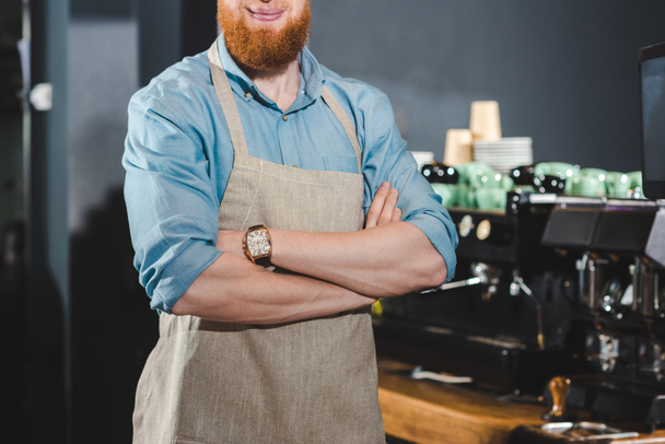 ritagliato colpo di barista barbuto sorridente in grembiule in piedi con le braccia incrociate in caffetteria
 - Foto, immagini