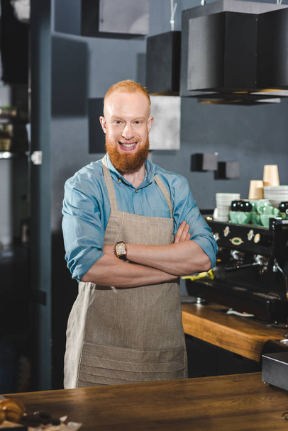 handsome young bearded barista standing with crossed arms and smiling at camera in coffee shop - Foto, Imagen