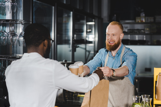 smiling bearded young barista giving paper bag and disposable coffee cups to african american buyer in cafe - Photo, Image