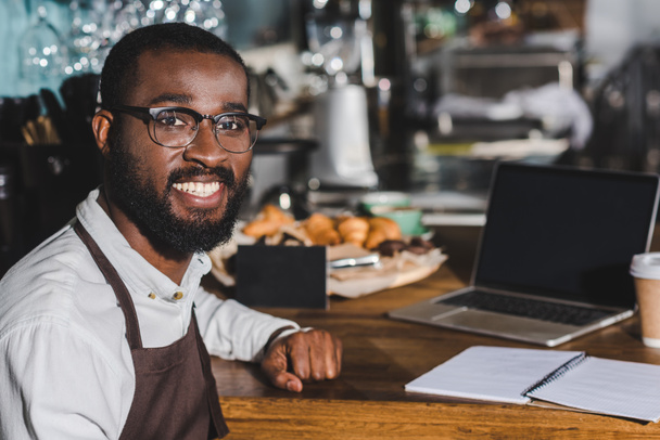 schöner junger afrikanisch-amerikanischer Barista mit Brille, der während seiner Arbeit im Café in die Kamera lächelt - Foto, Bild