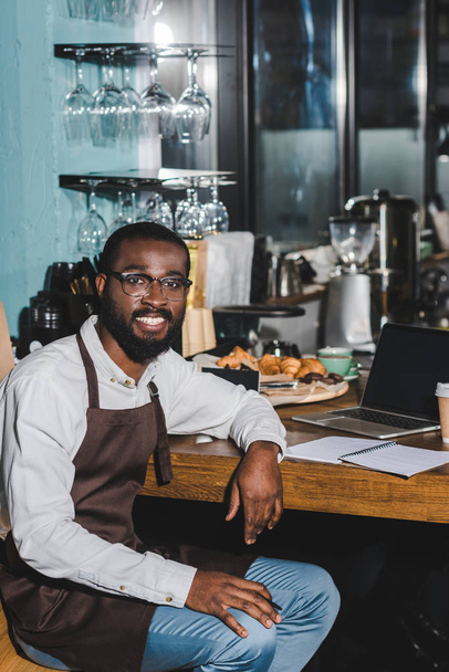 csinos fiatal afroamerikai barista a szemüvegek mosolyogva a kamera, miközben dolgozik a laptop a kávézóban - Fotó, kép