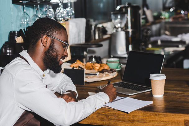 souriant jeune afro-américaine barista dans les lunettes prendre des notes et en utilisant un ordinateur portable avec écran blanc dans le café
 - Photo, image