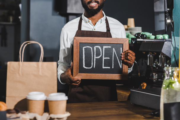 foto recortada de sonriente barista afroamericano sosteniendo cartel abierto en la cafetería
 - Foto, imagen