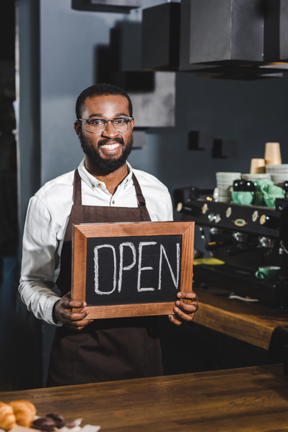 csinos fiatal afroamerikai barista a gazdaság jel megnyitása és mosolygós, a kamera a kávézóban szemüvegek - Fotó, kép
