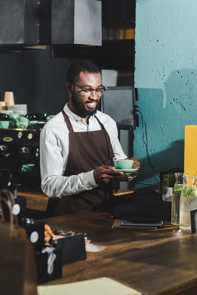 handsome smiling african american barista in eyeglasses holding cup of coffee in cafe - Photo, Image