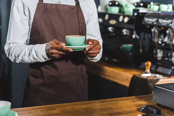tiro recortado de barista americano africano segurando xícara de cappuccino no café
 - Foto, Imagem