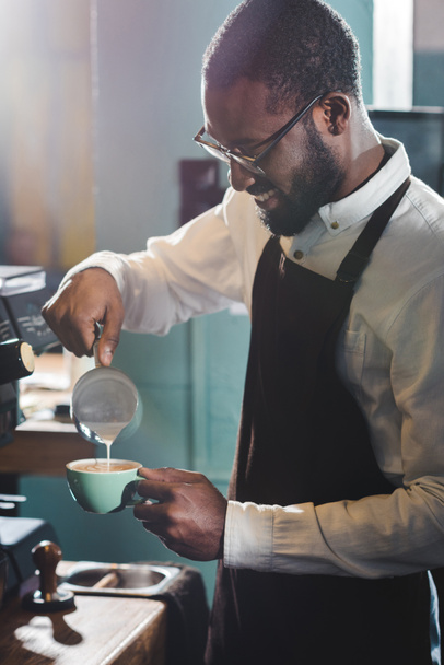 glimlachend jonge Afro-Amerikaanse barista in brillen gieten melk terwijl het maken van cappuccino - Foto, afbeelding