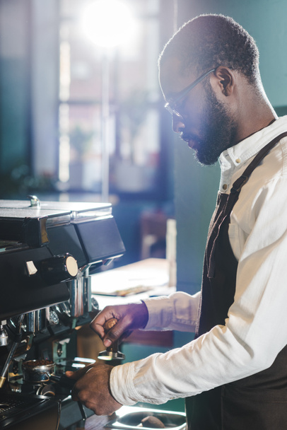 コーヒー マシンでコーヒーを準備する眼鏡の若いアフリカ系アメリカ人のバリスタの側面図 - 写真・画像