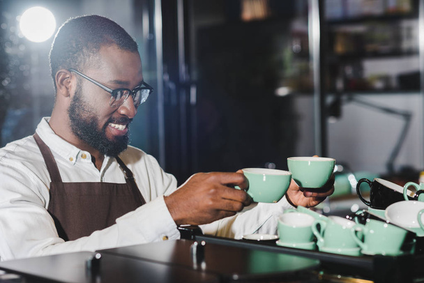 hymyilevä nuori afrikkalainen amerikkalainen barista silmälaseissa järjestämässä kuppeja kahvinkeittimen päälle
  - Valokuva, kuva