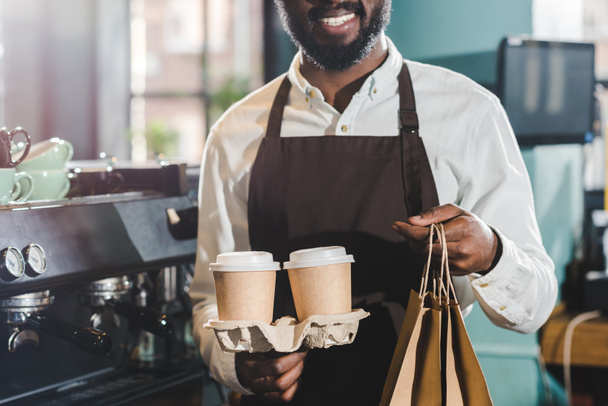 περικοπεί το πλάνο της χαμογελώντας αφρικανική αμερικανική barista κρατώντας χάρτινες τσάντες και φλιτζάνια μίας χρήσης καφέ σε καφετέρια - Φωτογραφία, εικόνα