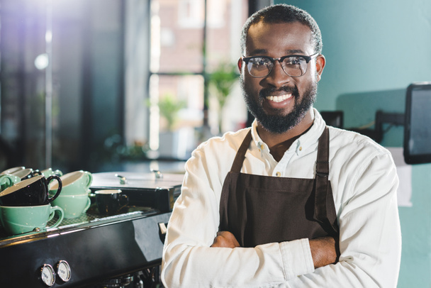 alegre africano americano barista en delantal y gafas de pie con los brazos cruzados y sonriendo a la cámara en la cafetería
 - Foto, imagen