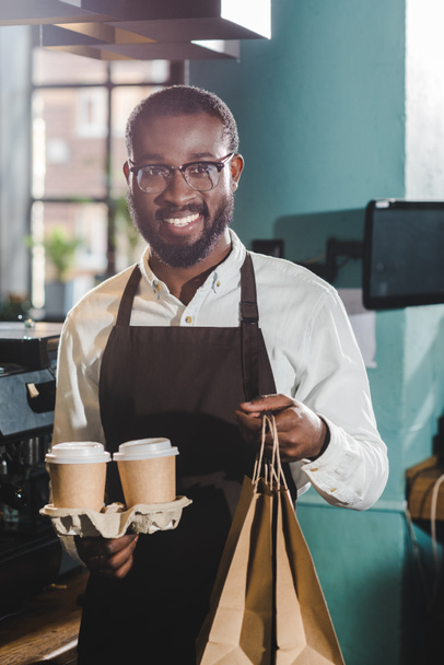 alegre joven africano americano barista celebración de papel tazas y bolsas en la cafetería
 - Foto, imagen