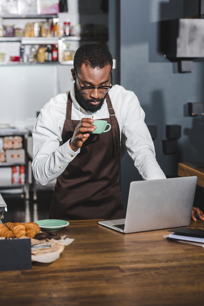 junger afrikanisch-amerikanischer Barista hält Tasse Kaffee in der Hand und benutzt Laptop im Café - Foto, Bild