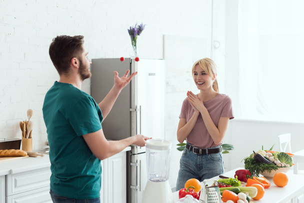 vegan boyfriend juggling with cherry tomatoes for happy girlfriend at kitchen - Photo, Image