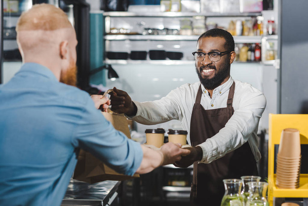 uśmiechający się młody Afroamerykanin barista dając kawy jednorazowe kubki i worek papierowy do klienta w kawiarni - Zdjęcie, obraz