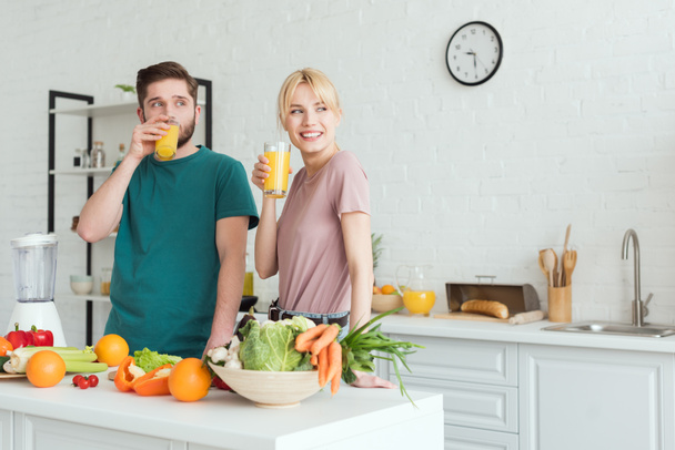 sorrindo casal de vegans bebendo suco fresco na cozinha e olhando para longe
 - Foto, Imagem