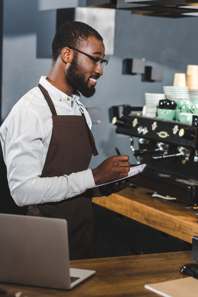 sorridente giovane barista afroamericano in occhiali prendere appunti mentre woeking in caffè
 - Foto, immagini