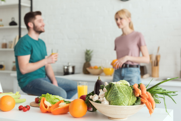 vegaani pari puhuu keittiössä hedelmiä ja vihanneksia etualalla
 - Valokuva, kuva