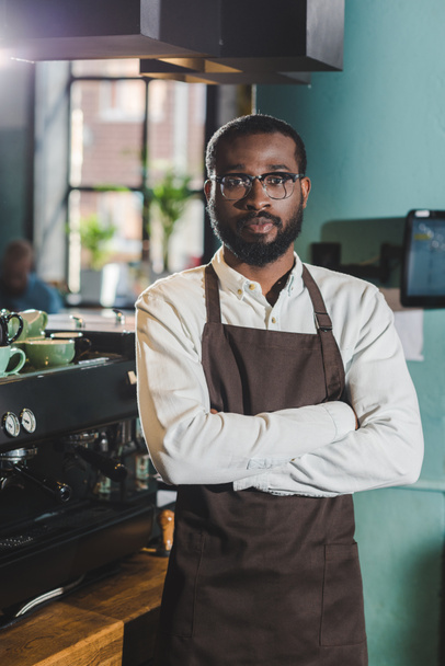handsome african american barista standing with crossed arms and looking at camera in coffee shop  - Fotoğraf, Görsel