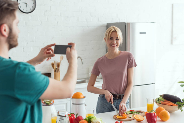 частичный вид человека, фотографирующего улыбающуюся подругу, готовящую дома на кухне, концепция веганского образа жизни
 - Фото, изображение