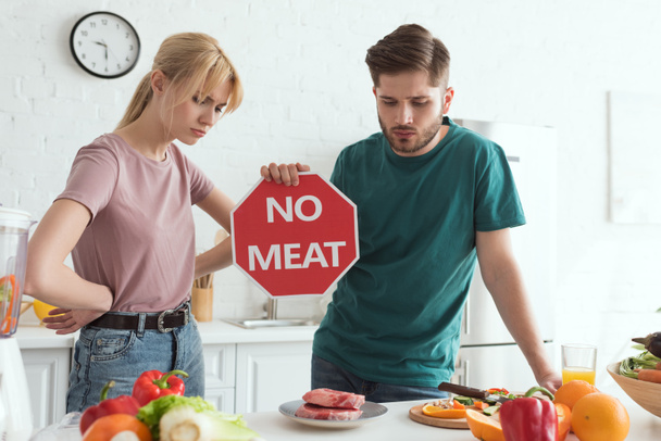 vegan couple with no meat sign at table with raw meat and vegetables in kitchen at home - Photo, Image