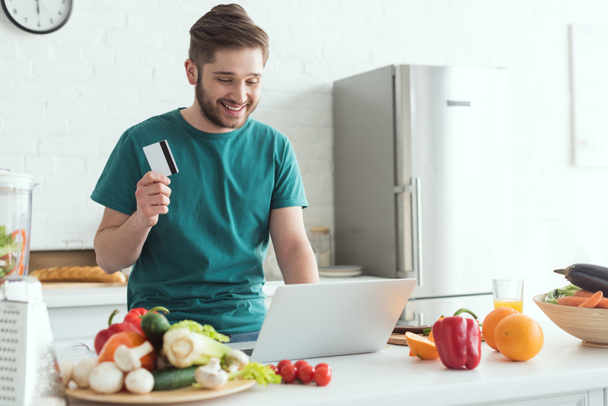 mosolygó ember hitelkártya és laptop adásvétel online otthon a konyhában - Fotó, kép