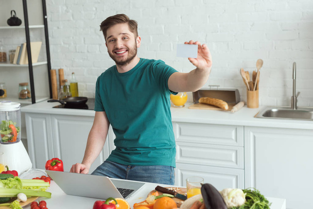 hombre sonriente mostrando la tarjeta de crédito en la mesa con el ordenador portátil en la cocina en casa
 - Foto, imagen