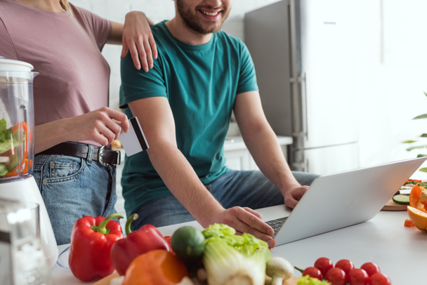 vista parcial de pareja vegana con ordenador portátil de compras en línea en la cocina en casa
 - Foto, imagen