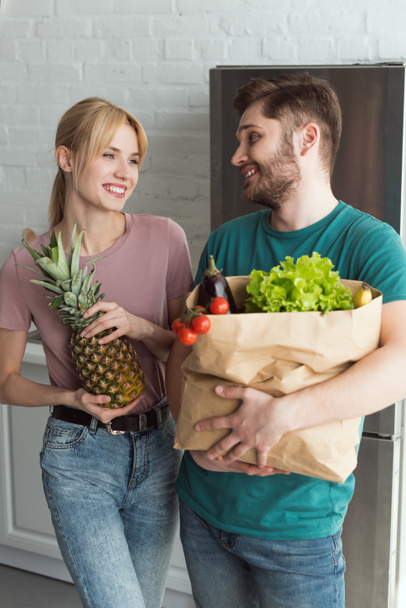 紙袋の自宅のキッチンで新鮮な野菜と笑顔のビーガン カップル - 写真・画像