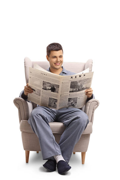 Young guy in pajamas sitting in an armchair and reading a newspaper isolated on white background - Foto, Imagem