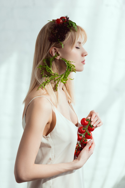Πλάγια όψη της νεαρής γυναίκας με ντοματίνια στα χέρια, στεφάνι φτιαγμένο από φρέσκα λαχανικά και σκουλαρίκι φιαγμένος από φρέσκια ρόκα, έννοια τρόπου ζωής για χορτοφάγους - Φωτογραφία, εικόνα