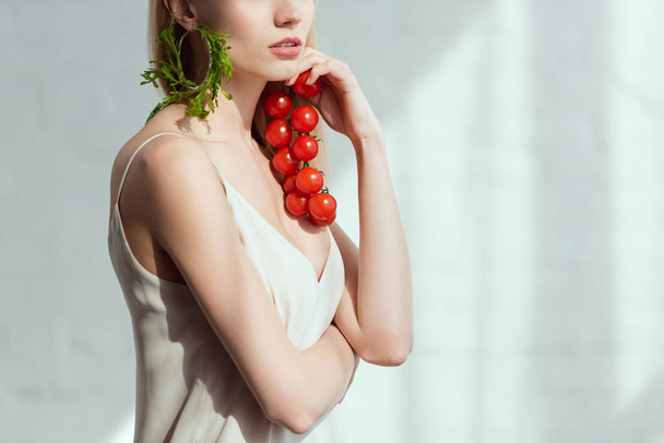 partial view of woman with cherry tomatoes in hand and earring made of fresh arugula, vegan lifestyle concept - Photo, Image