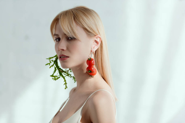 thoughtful woman in dress with earrings made of fresh arugula and cherry tomatoes, vegan lifestyle concept - Photo, Image