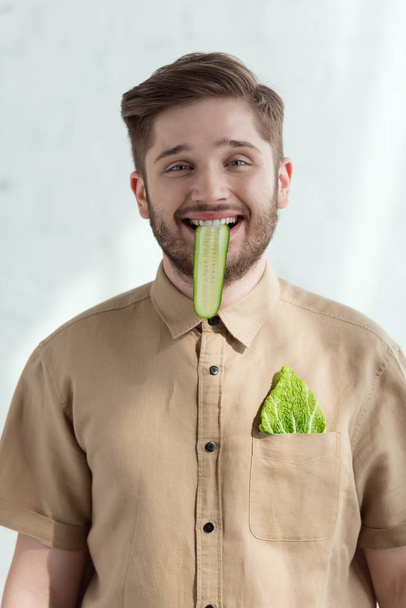 Portret van glimlachende man met komkommer segment in mond en Savoye kool blad in zak, veganistische levensstijl concept  - Foto, afbeelding
