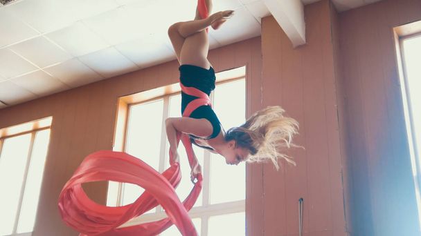 Приваблива дівчина повітряна гімнастка звисає догори ногами і махає повітряним шовком
 - Фото, зображення